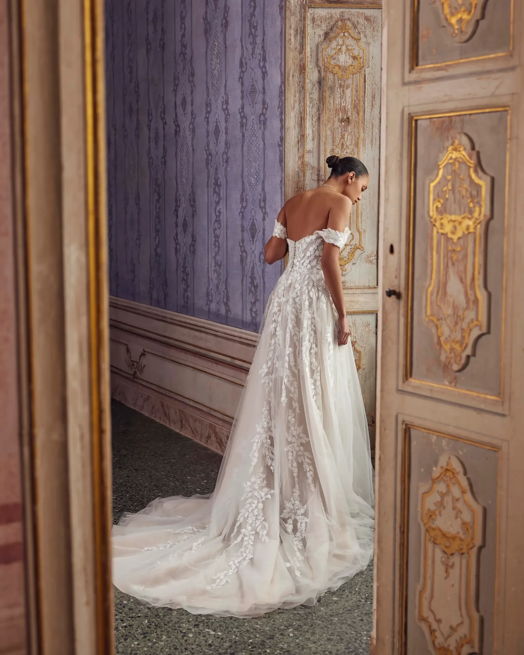 A woman in a white wedding dress stands with her back to the camera, looking down, in an ornate room with purple walls and intricate doors.