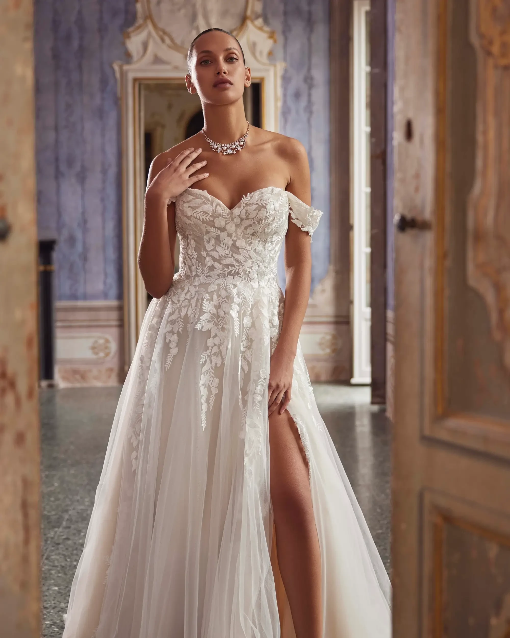 A model wearing an elegant off-the-shoulder wedding gown with floral designs and a thigh-high slit, posing in a luxurious setting.