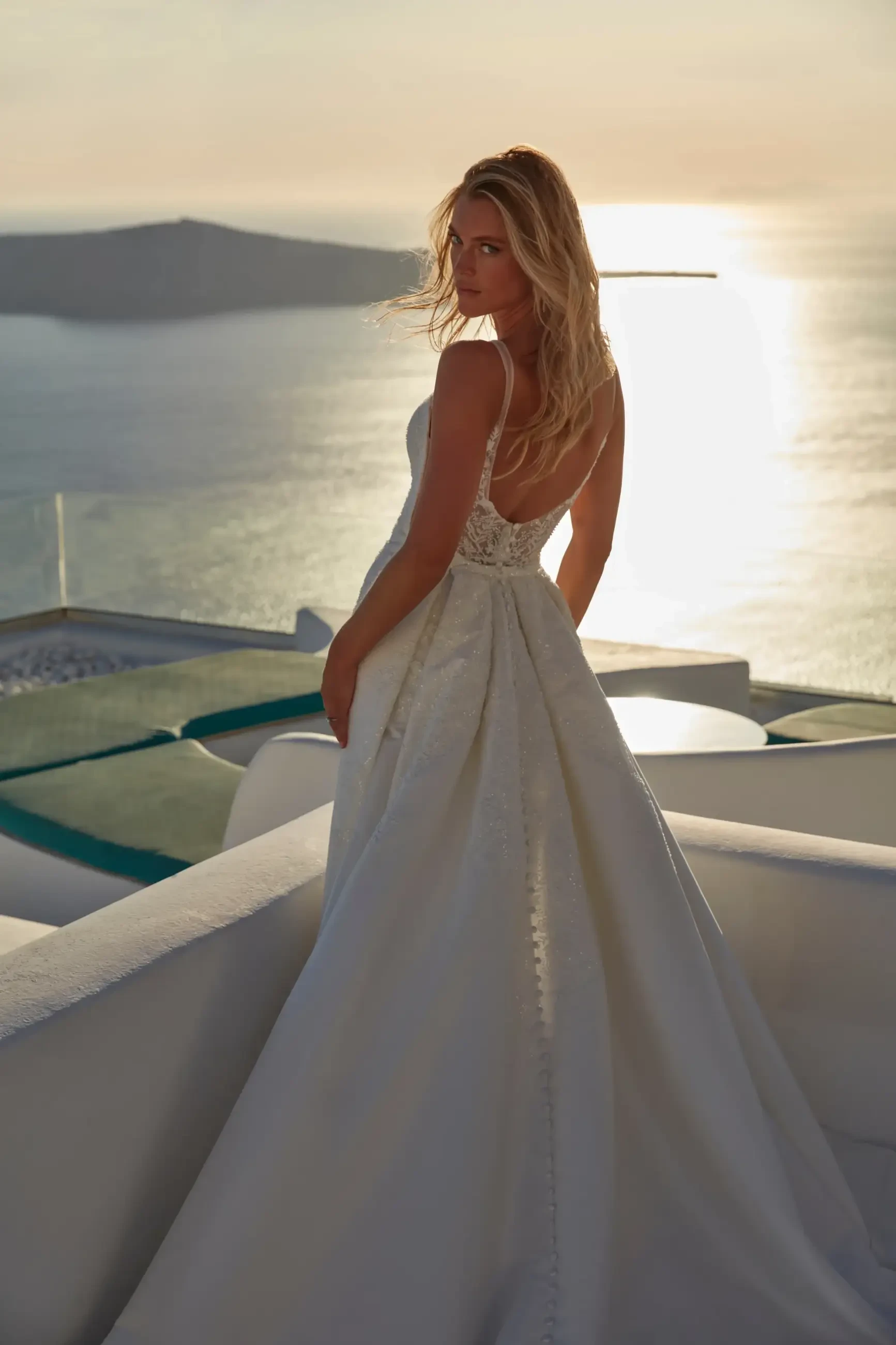 A woman in a flowing white gown stands with her back to the camera, looking out over a body of water during sunset.