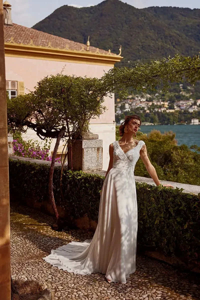 A woman in a beautiful wedding dress posing outdoors near lush greenery with mountains and water in the background.