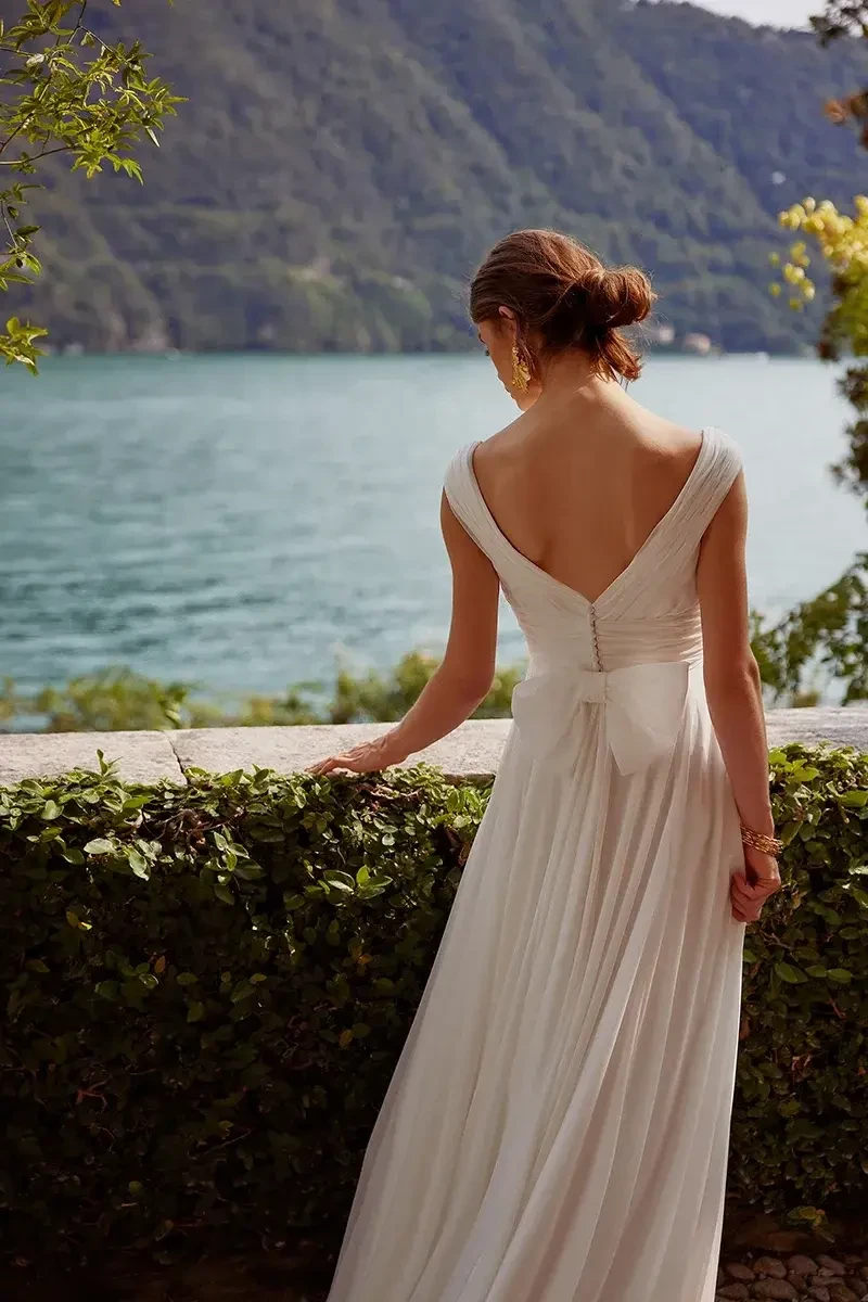 A woman in a white dress stands by a body of water, looking towards the landscape.
