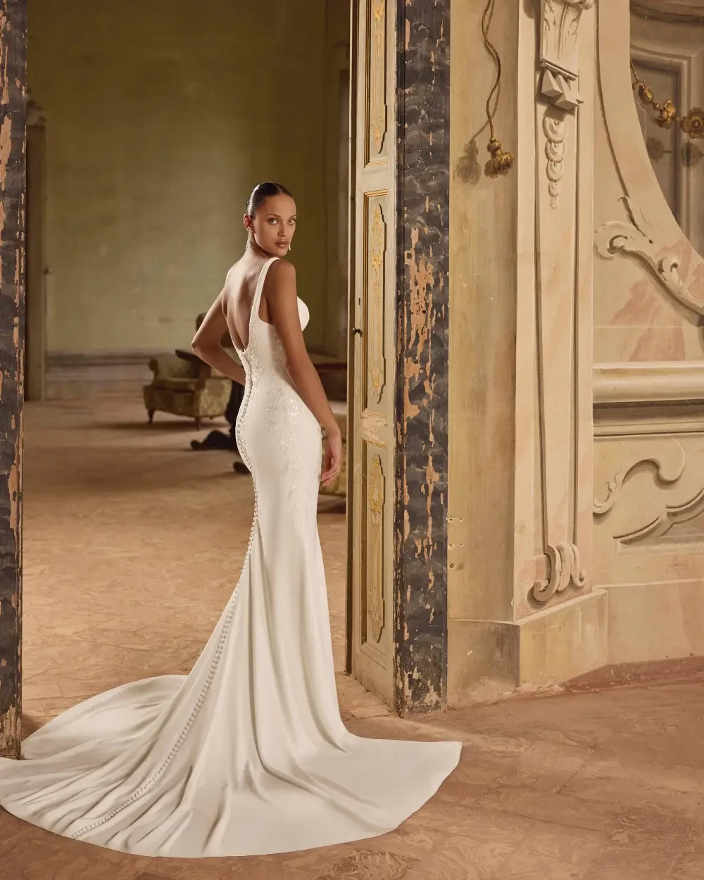 A woman in a sleek, fitted white gown with a train, standing gracefully in a vintage-style room with ornate decor.