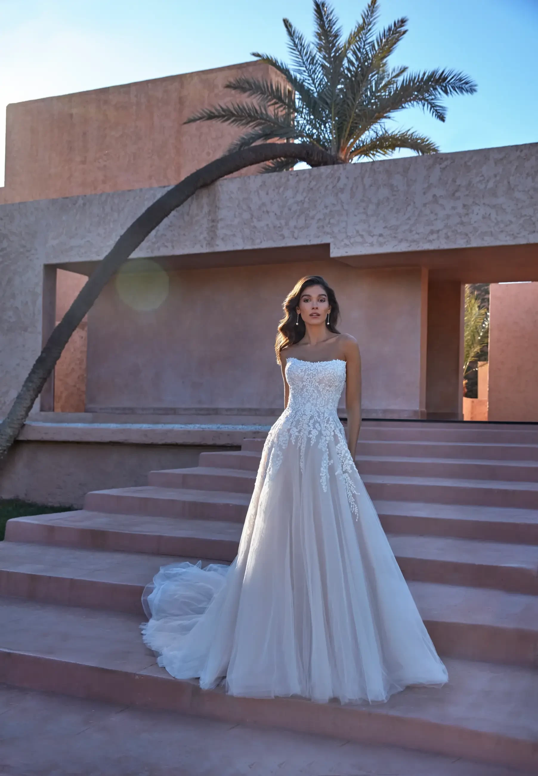 A woman in a wedding dress stands on steps with a palm tree in the background and geometric architecture.