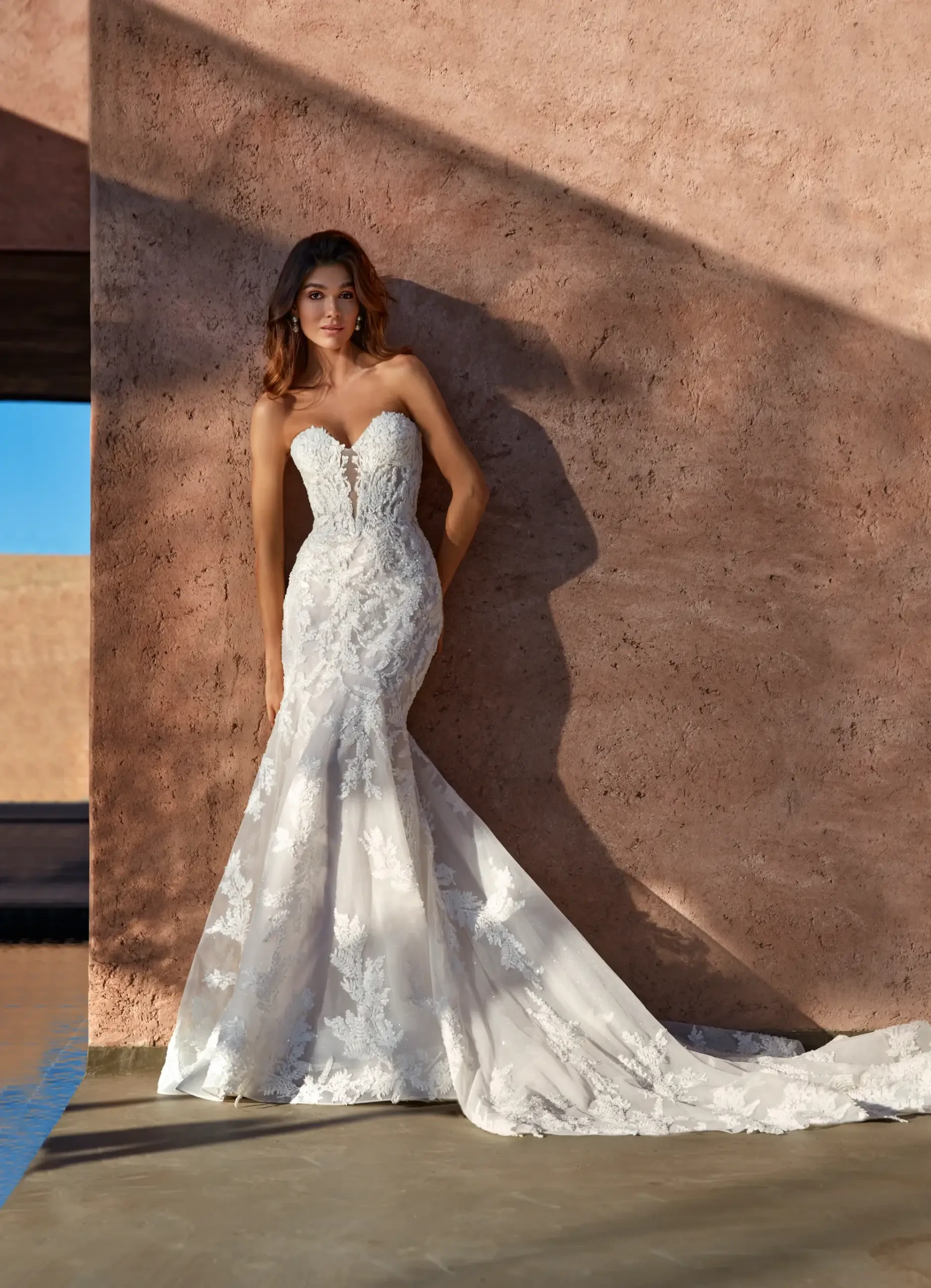 A woman wearing a strapless, lace wedding dress that flares out at the bottom, standing against a textured wall with soft lighting.