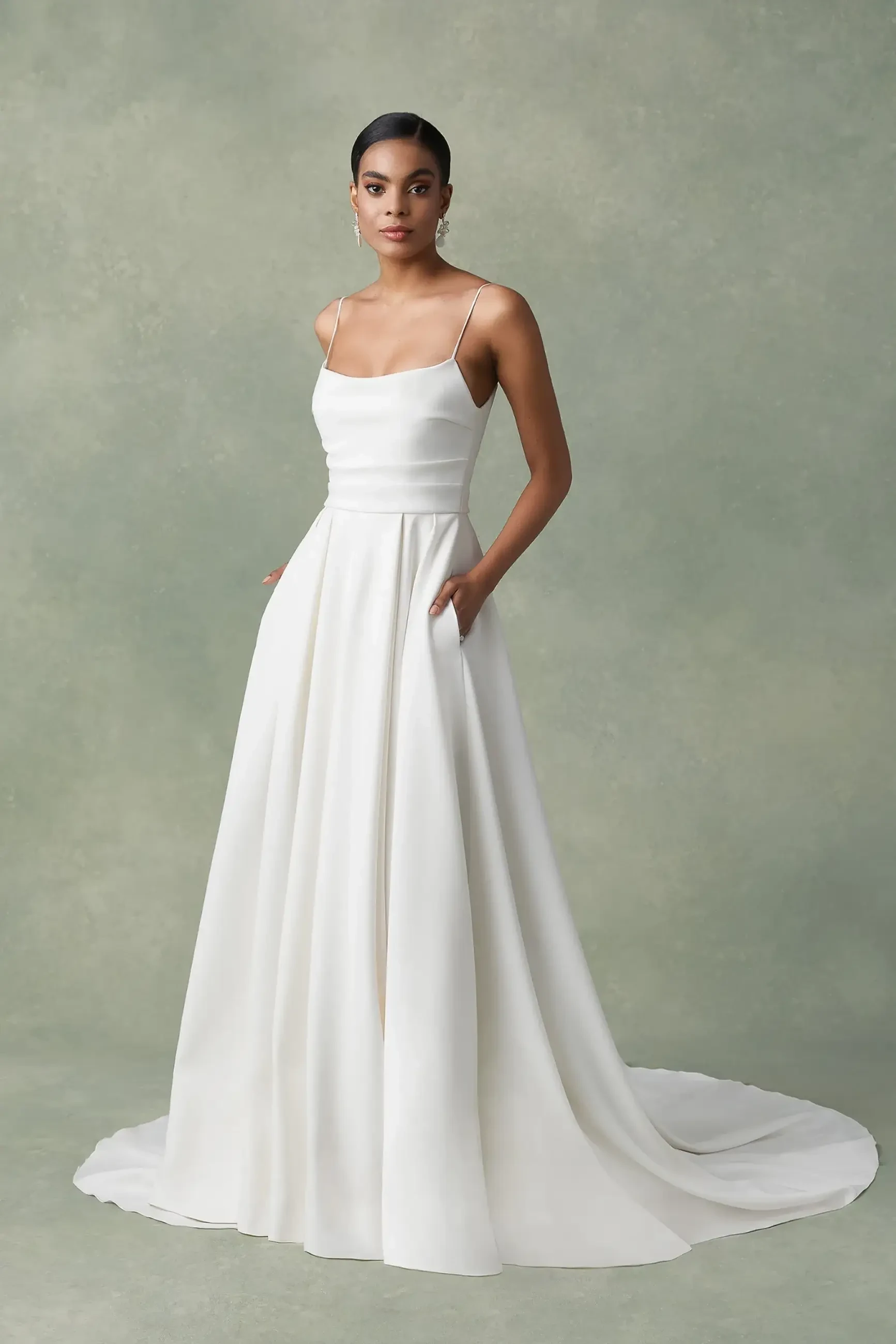 A woman stands in a simple, elegant white gown with a fitted bodice and flowing skirt, featuring thin straps and pockets. The background is softly textured in muted tones.