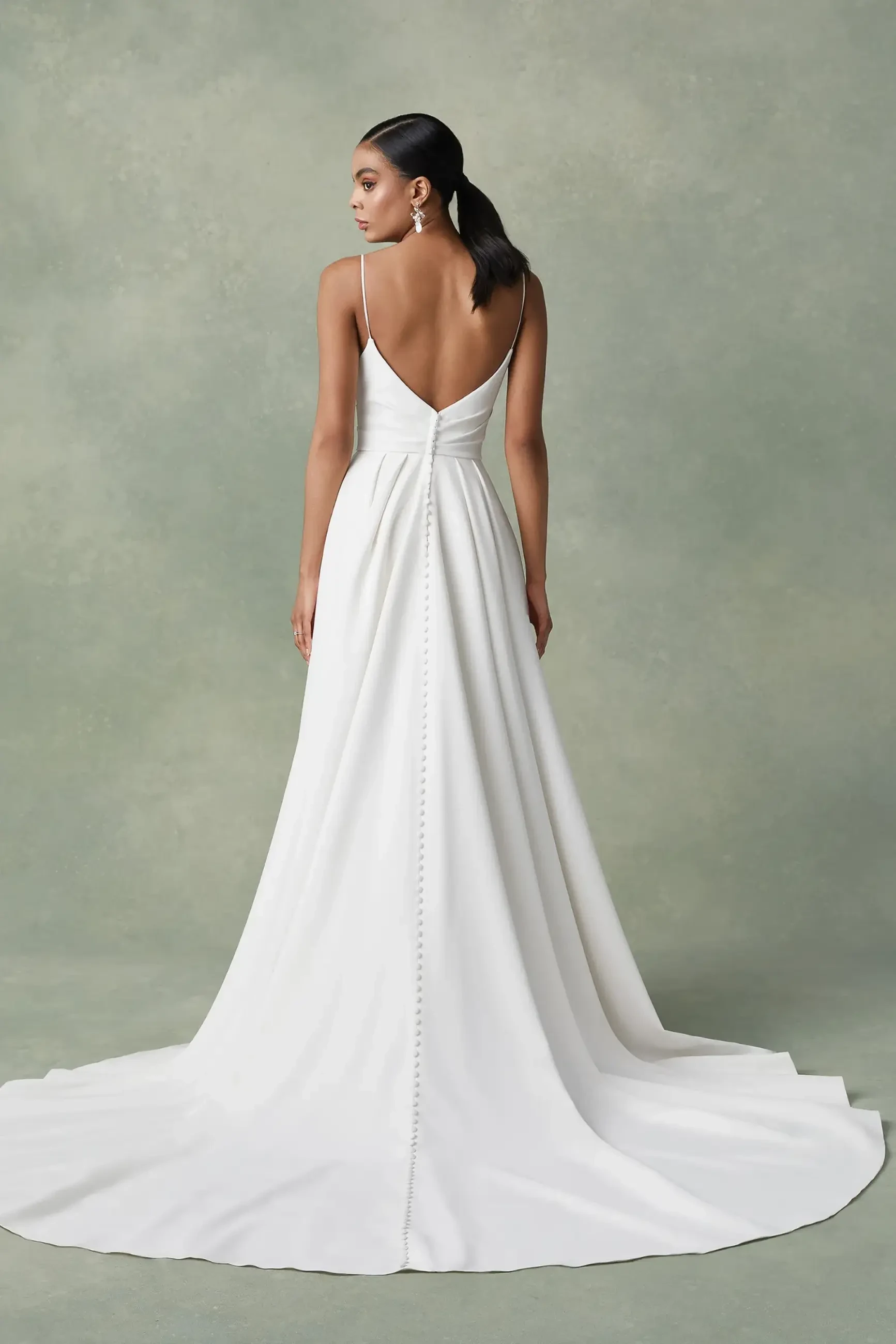 A model wearing a white wedding dress with a fitted bodice and a flowing train, viewed from the back. The dress features thin straps and a keyhole detail at the back, with buttons running down the skirt.