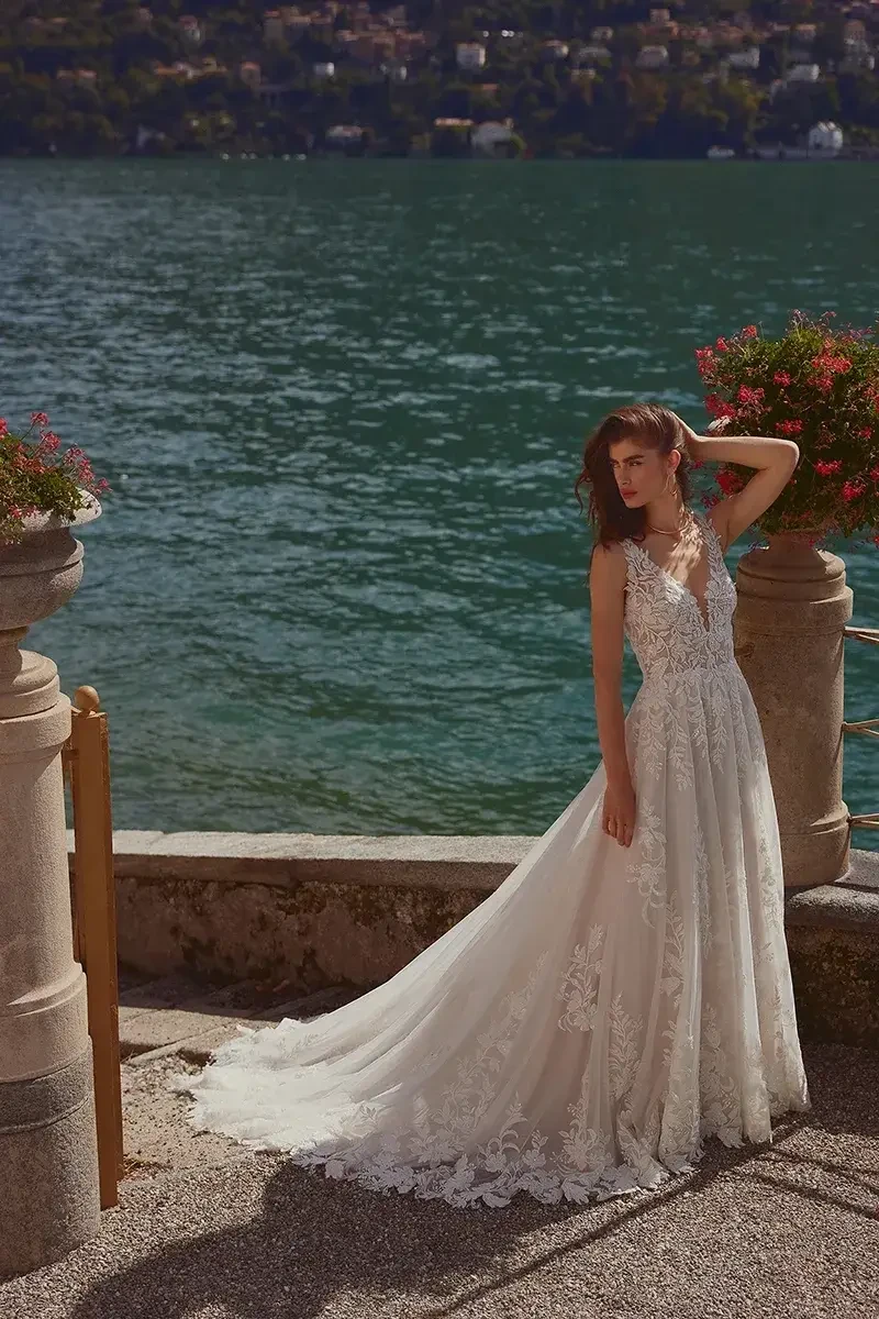 A woman in a flowing, elegant bridal gown stands by a body of water, with decorative planters and a scenic background.