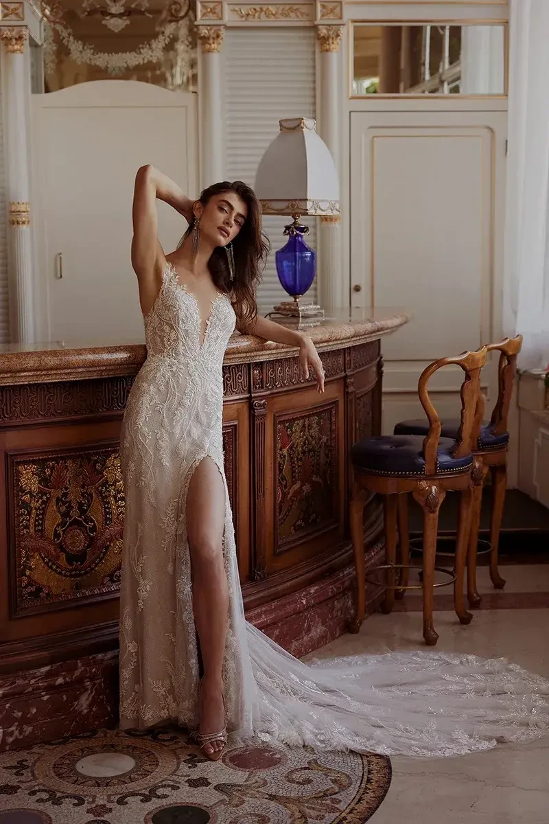 A woman in an elegant wedding dress with intricate lace details poses beside a beautifully decorated bar. The dress features a thigh-high slit, and the setting is brightly lit with ornate decor.