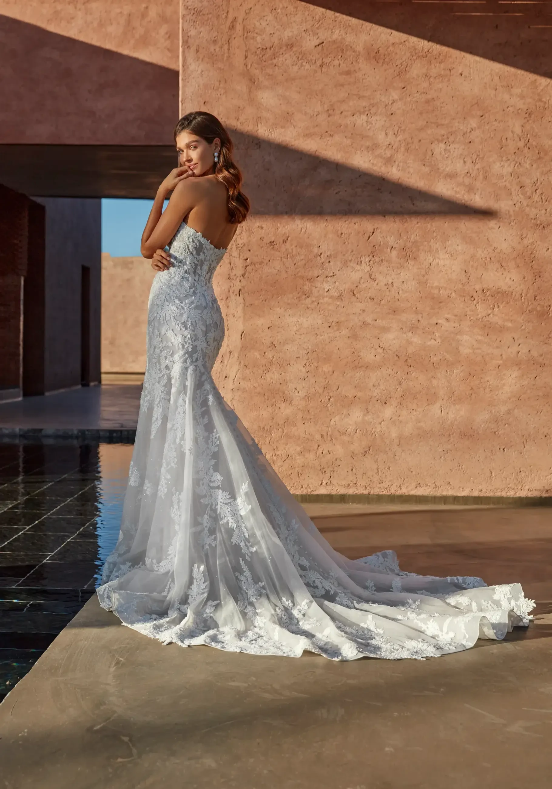 A woman wearing a white lace wedding dress stands gracefully, showcasing the gown's train and intricate details, against a warm-toned architectural backdrop.