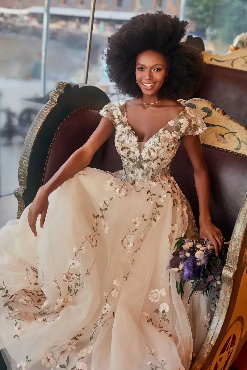 A woman with an afro hairstyle is sitting on a decorative chair, wearing a floral embroidered gown and holding a bouquet of flowers.